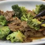 Beef and broccoli stir fry served in a white bowl on a wooden surface, with chopsticks lifting a piece of broccoli