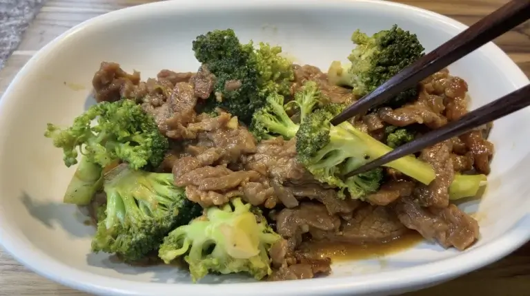 Beef and broccoli stir fry served in a white bowl on a wooden surface, with chopsticks lifting a piece of broccoli